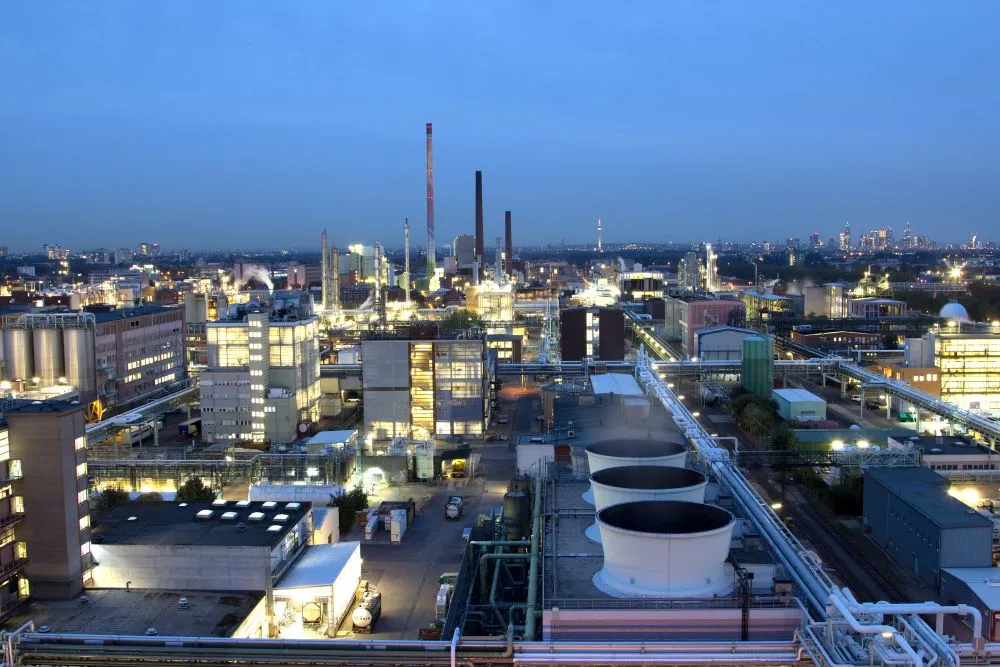 Industriepark mit beleuchteten Fabrikgebäuden, hohen Schornsteinen und großen Lüftungstürmen bei Dämmerung.