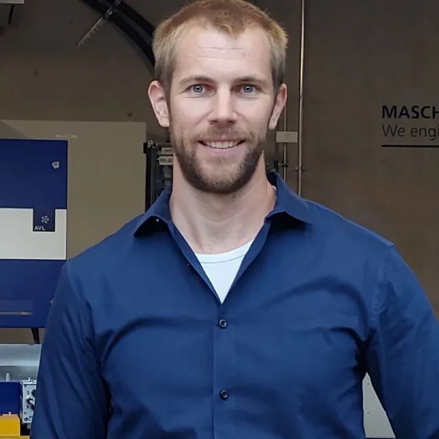 Mann mit kurzem blondem Haar und Bart trägt blaues Hemd und steht vor einer Werkstattwand mit Maschinen und Regalen