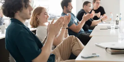 Mehrere Personen sitzen an einem Tisch und klatschen mit sichtbaren Händen und Oberkörpern.