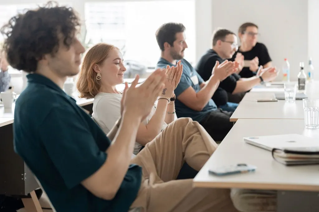 Mehrere Personen sitzen an einem Tisch und klatschen mit sichtbaren Händen und Oberkörpern.