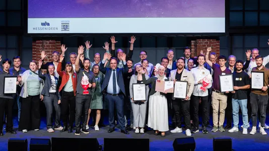Gruppe von Menschen auf Bühne vor Backsteinwand mit Urkunden und Pokalen, einige heben die Hände