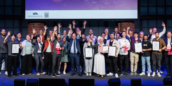 Gruppe von Menschen auf Bühne vor Backsteinwand mit Urkunden und Pokalen, einige heben die Hände