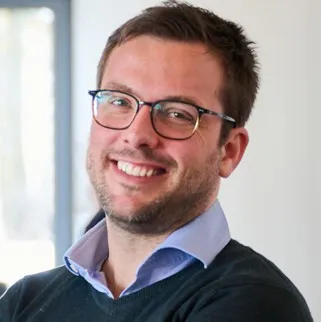 Mann mit kurz geschnittenem Haar, hellblauem Hemd und dunklem Pullover vor hellem Hintergrund