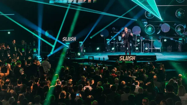 Mann auf Bühne spricht vor großem Publikum bei Slush-Event, umgeben von bunten Lichtstrahlen und Bühnenbild mit Slush-Logo.