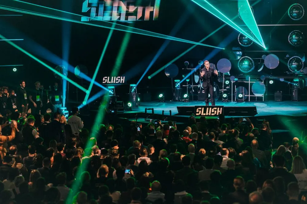 Mann auf Bühne spricht vor großem Publikum bei Slush-Event, umgeben von bunten Lichtstrahlen und Bühnenbild mit Slush-Logo.