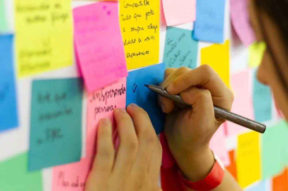 Person schreibt mit Stift auf einen blauen Haftnotizzettel an einer Wand mit vielen bunten Haftnotizen