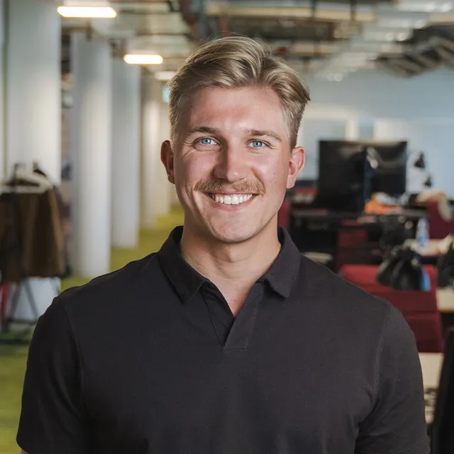 Mann mit blondem Haar und schwarzem Poloshirt in einem Büro mit Regalen und Computern im Hintergrund