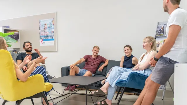 Fünf Personen sitzen und stehen in einem modernen Raum mit Sesseln und Sofas und unterhalten sich entspannt