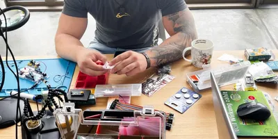 Oberkörper eines Mannes mit Tattoos, der an einem Tisch mit Elektronikbauteilen und Werkzeugen sitzt