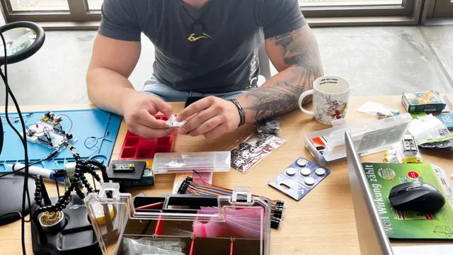 Oberkörper eines Mannes mit Tattoos, der an einem Tisch mit Elektronikbauteilen und Werkzeugen sitzt