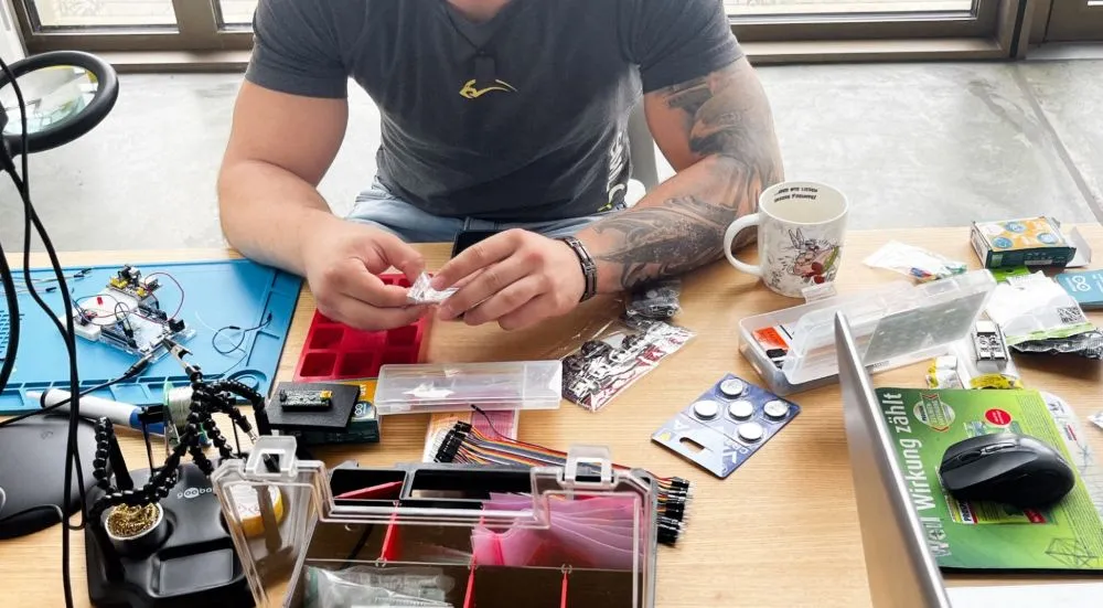 Oberkörper eines Mannes mit Tattoos, der an einem Tisch mit Elektronikbauteilen und Werkzeugen sitzt
