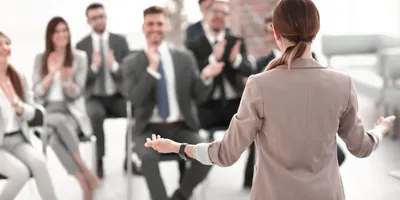 Frau mit Zopf und beigem Blazer spricht vor sitzendem Publikum, das applaudiert