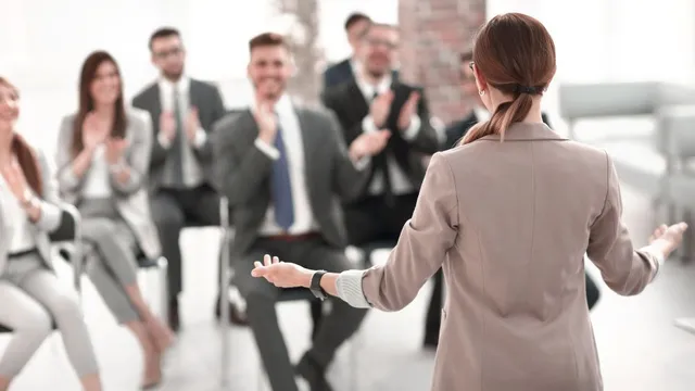 Frau mit Zopf und beigem Blazer spricht vor sitzendem Publikum, das applaudiert
