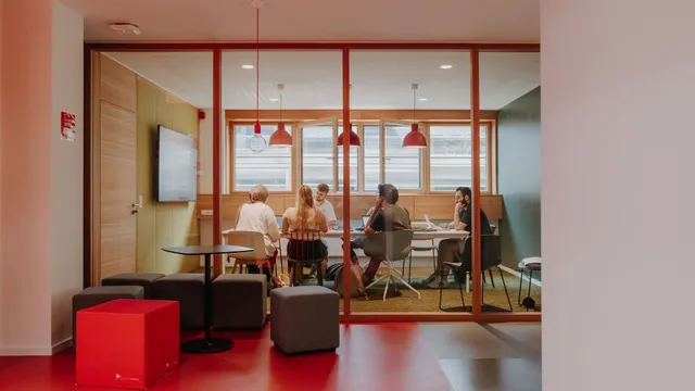 Blick durch eine Glaswand auf einen Besprechungsraum mit fünf Personen an einem Tisch und mehreren Sitzwürfeln im Vordergrund