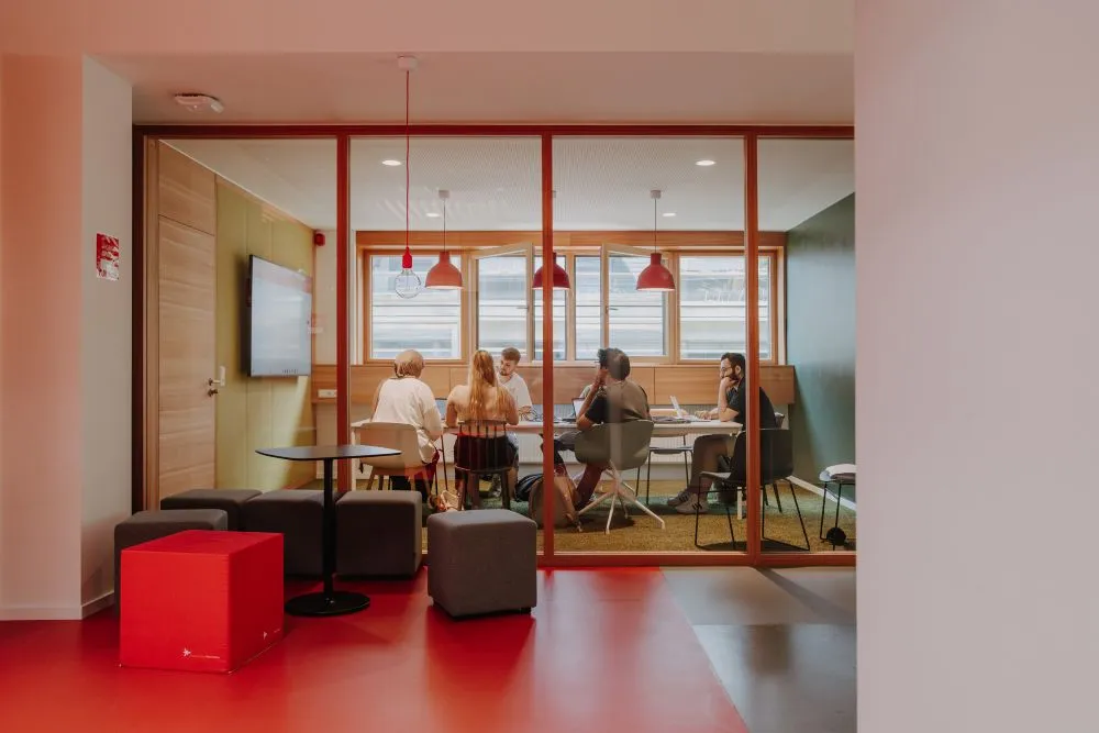 Blick durch eine Glaswand auf einen Besprechungsraum mit fünf Personen an einem Tisch und mehreren Sitzwürfeln im Vordergrund