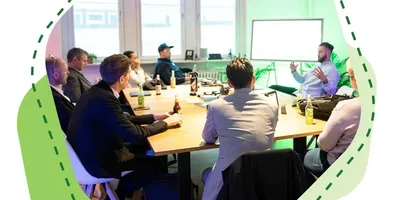 Mehrere Personen sitzen an einem Tisch und hören einem Mann zu, der vor einem Whiteboard spricht