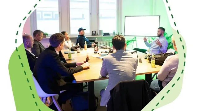Mehrere Personen sitzen an einem Tisch und hören einem Mann zu, der vor einem Whiteboard spricht