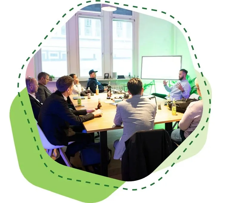 Mehrere Personen sitzen an einem Tisch und hören einem Mann zu, der vor einem Whiteboard spricht