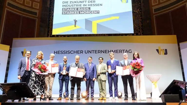 Personengruppe auf Bühne beim Hessischen Unternehmertag, einige halten Urkunden, zwei Frauen tragen Blumensträuße, Hintergrund mit Veranstaltungstitel und Slogan.