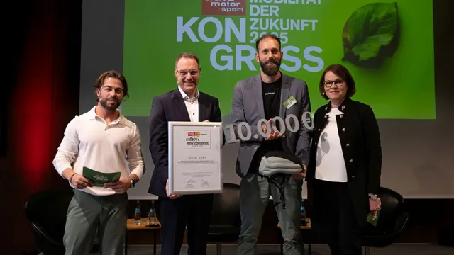 Vier Personen stehen vor einer grünen Leinwand mit dem Text 'auto motor sport MOBILITÄT DER ZUKUNFT 2025 KONGRESS'. Eine Person hält eine Urkunde, eine andere ein Schild mit '10.000 €.