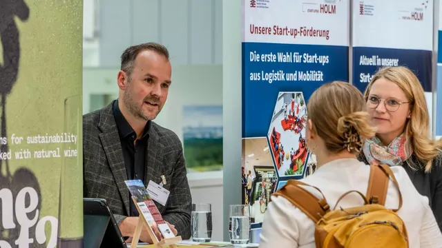 Zwei Frauen mit Rucksack und Schal sprechen mit einem Mann in kariertem Sakko an einem Messestand mit Bannern zur Start-up-Förderung von HOLM