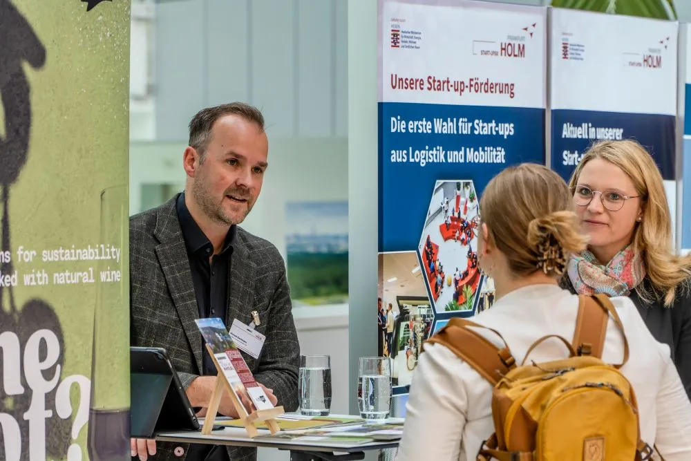 Zwei Frauen mit Rucksack und Schal sprechen mit einem Mann in kariertem Sakko an einem Messestand mit Bannern zur Start-up-Förderung von HOLM