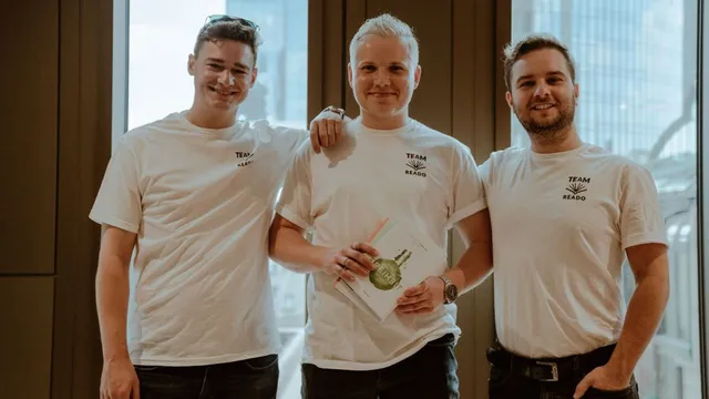 Drei Männer stehen nebeneinander vor einem Fenster, tragen weiße T-Shirts mit der Aufschrift 'TEAM READO', der mittlere hält ein Buch mit einer Medaille auf dem Cover
