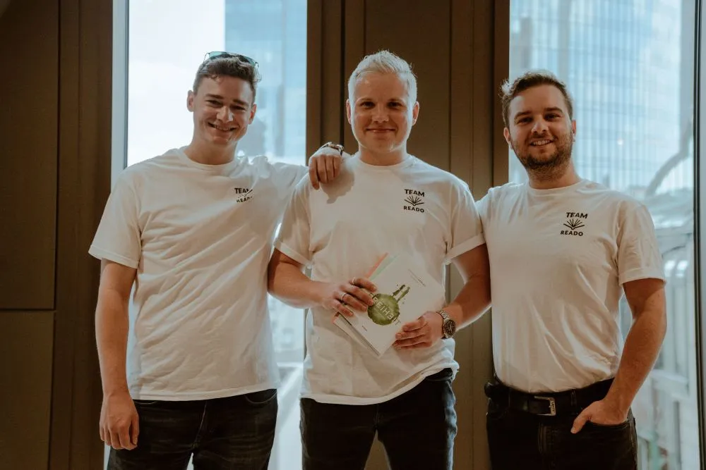 Drei Männer stehen nebeneinander vor einem Fenster, tragen weiße T-Shirts mit der Aufschrift 'TEAM READO', der mittlere hält ein Buch mit einer Medaille auf dem Cover