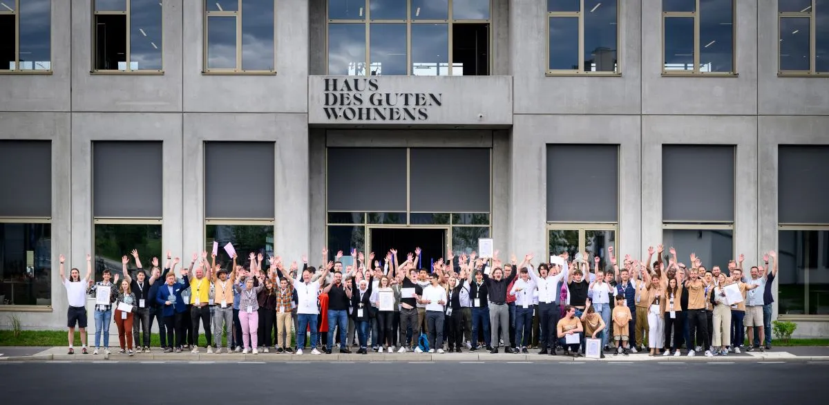 Große Gruppe von Menschen vor dem Gebäude mit Schild 'Haus des guten Wohnens', alle winken und lächeln