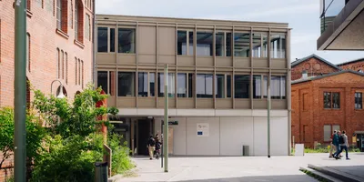 Moderner, hellgrauer Büroanbau zwischen roten Backsteingebäuden, Personen gehen auf dem gepflasterten Hof.