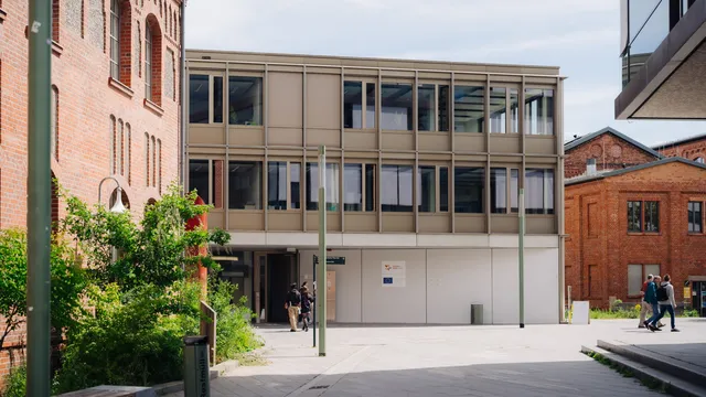 Moderner, hellgrauer Büroanbau zwischen roten Backsteingebäuden, Personen gehen auf dem gepflasterten Hof.
