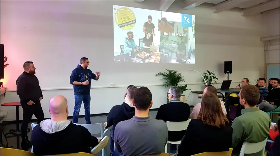 Mehrere Personen sitzen in einem Raum und hören einem Mann zu, der vor einer Wand mit einer Präsentation steht, auf der 'Social Pizza' zu lesen ist.