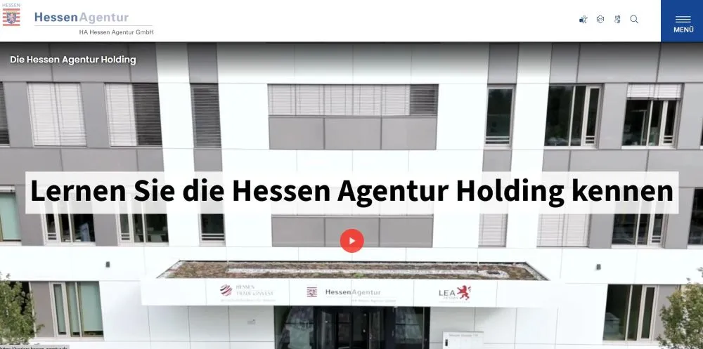 Fassade eines modernen Bürogebäudes mit großen Fenstern und Textbanner 'Lernen Sie die Hessen Agentur Holding kennen' in schwarzer Schrift auf weißem Hintergrund.