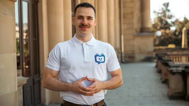 Mann mit Schnurrbart in weißem Poloshirt mit blauem Logo steht vor einem historischen Gebäude und hält die Hände vor dem Bauch zusammen