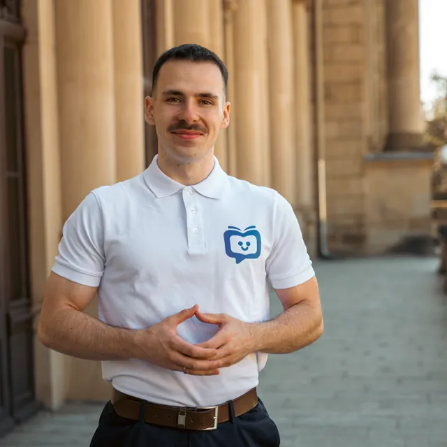 Mann mit Schnurrbart in weißem Poloshirt mit blauem Logo steht vor einem historischen Gebäude und hält die Hände vor dem Bauch zusammen