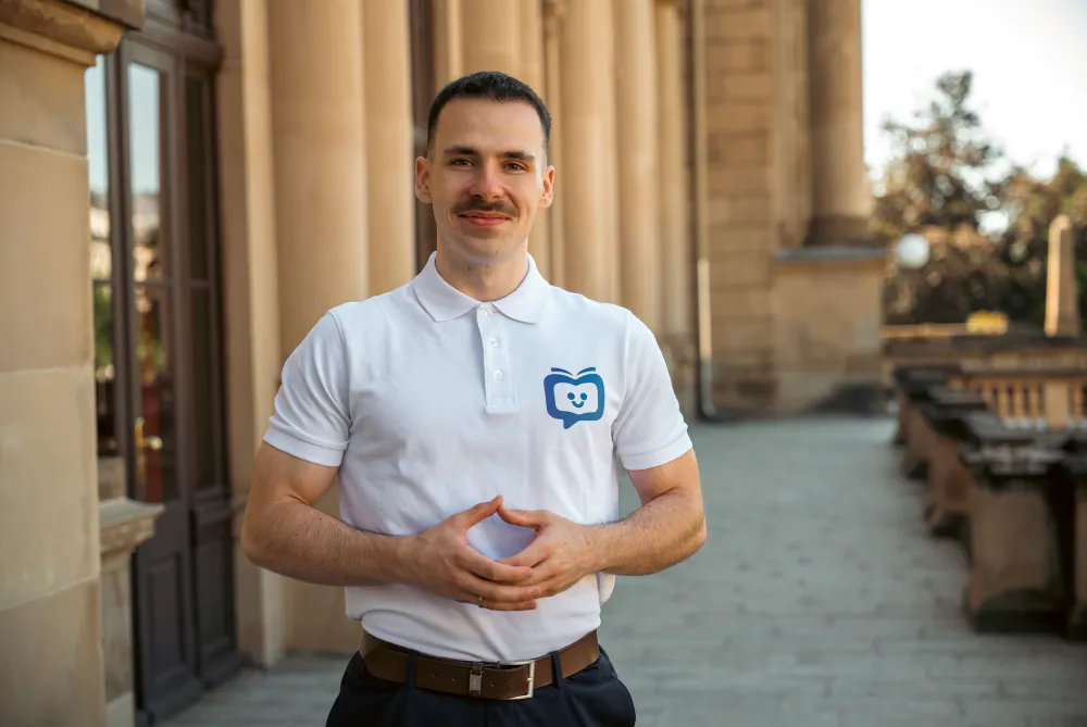 Mann mit Schnurrbart in weißem Poloshirt mit blauem Logo steht vor einem historischen Gebäude und hält die Hände vor dem Bauch zusammen