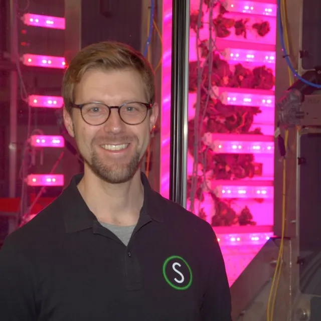 Mann mit schwarzem Poloshirt mit grün-weißem Logo vor vertikalen Pflanzregalen mit pinkfarbener Beleuchtung