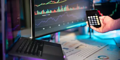 Hand hält Taschenrechner vor zwei Bildschirmen mit Finanzdiagrammen und Laptop