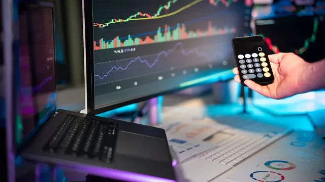 Hand hält Taschenrechner vor zwei Bildschirmen mit Finanzdiagrammen und Laptop
