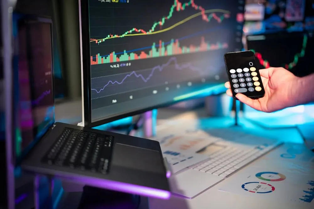 Hand hält Taschenrechner vor zwei Bildschirmen mit Finanzdiagrammen und Laptop