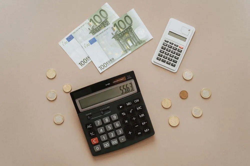 Zwei Taschenrechner, drei 100-Euro-Scheine und mehrere Euro-Münzen auf hellem Untergrund