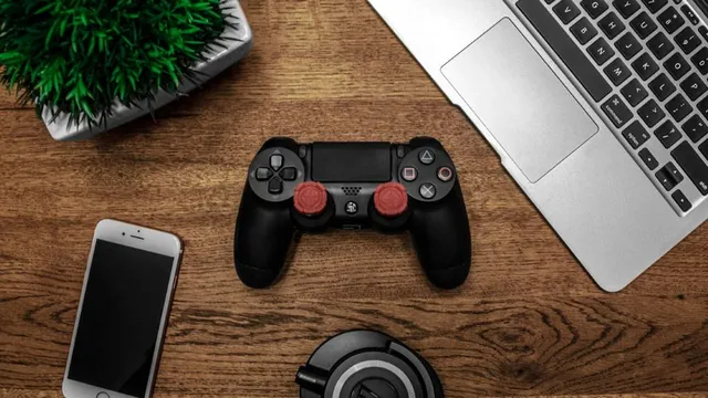 Schwarzer PlayStation-Controller mit roten Daumensticks, Smartphone, Laptop und Kopfhörern auf einem Holztisch.