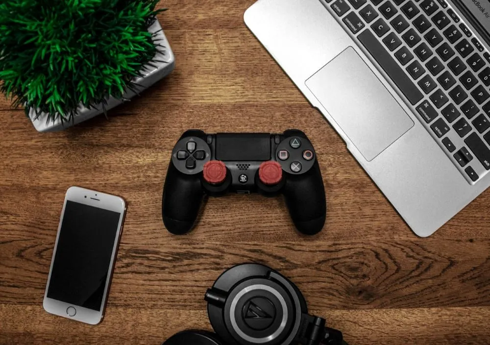 Schwarzer PlayStation-Controller mit roten Daumensticks, Smartphone, Laptop und Kopfhörern auf einem Holztisch.