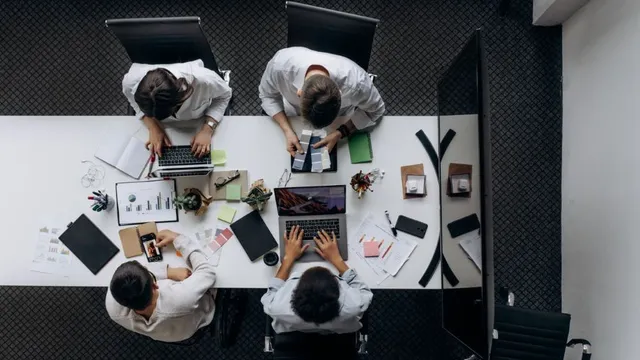 Vier Personen sitzen an einem Tisch und arbeiten mit Laptop, Smartphone und Unterlagen