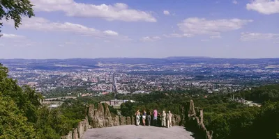Blick von einer Aussichtsplattform mit Felsen und mehreren Personen auf eine Stadt in einem Tal unter blauem Himmel mit Wolken