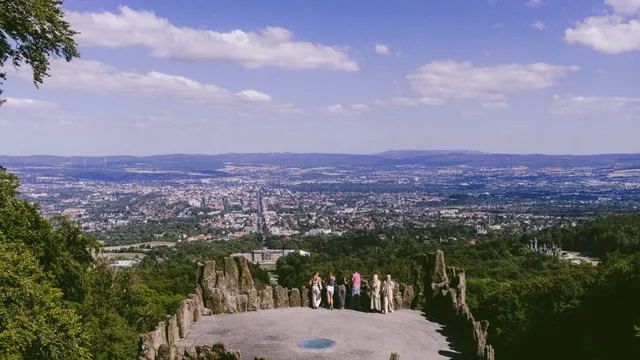 Blick von einer Aussichtsplattform mit Felsen und mehreren Personen auf eine Stadt in einem Tal unter blauem Himmel mit Wolken