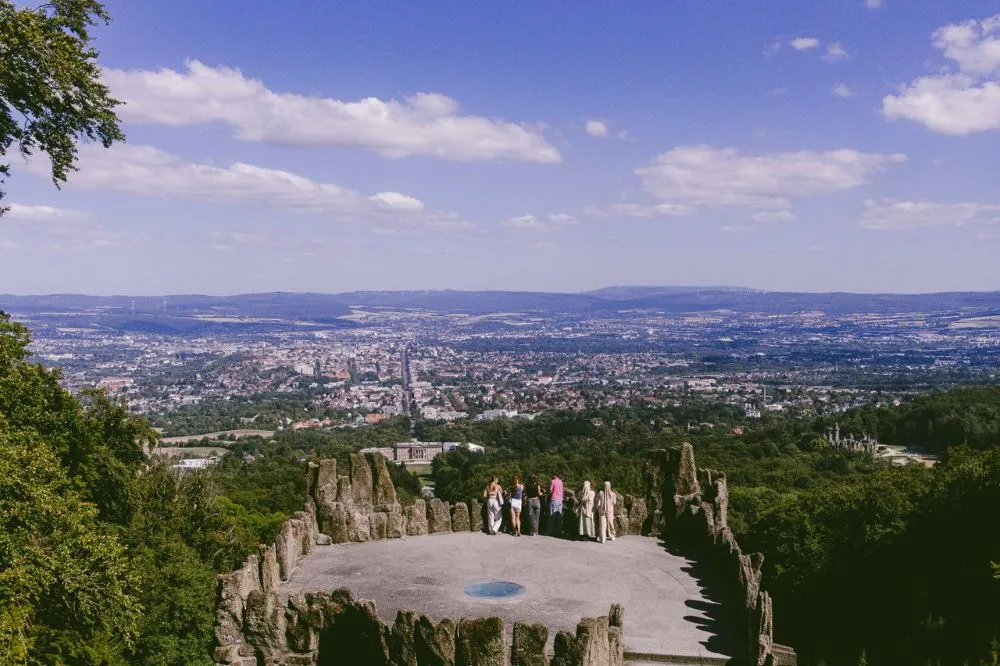 Blick von einer Aussichtsplattform mit Felsen und mehreren Personen auf eine Stadt in einem Tal unter blauem Himmel mit Wolken