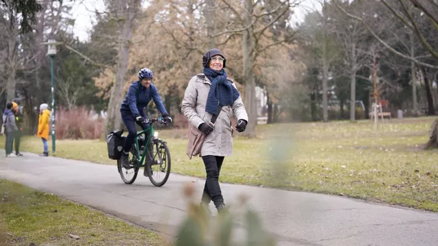 Person mit Schal und Mantel geht auf einem Weg in einem Park, im Hintergrund fährt eine Person mit Fahrrad