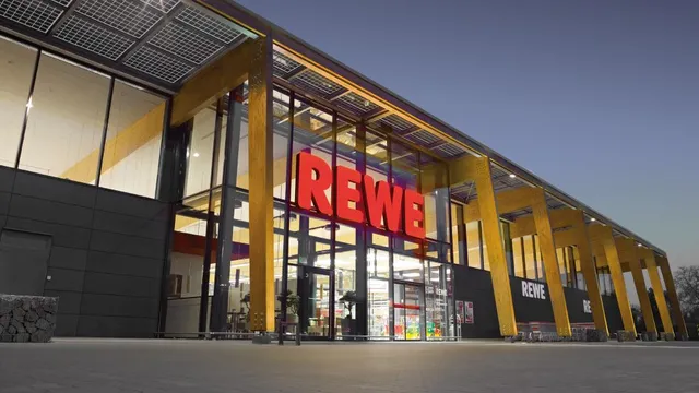 Eingang eines REWE-Supermarkts mit großem roten Schriftzug über Glastüren und moderner Holzfassade