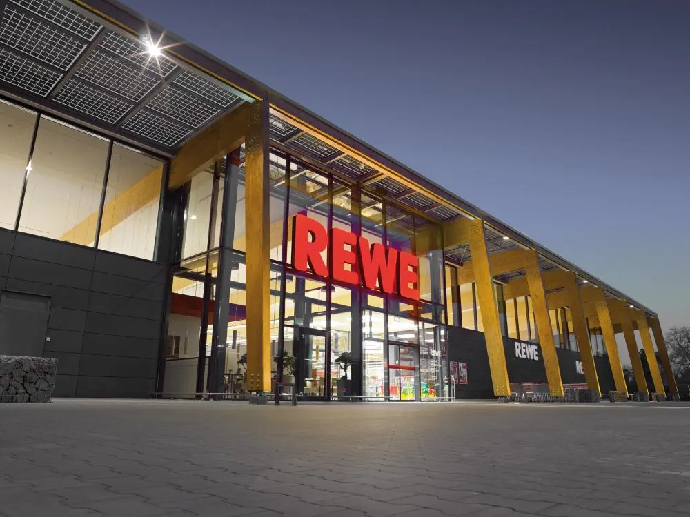 Eingang eines REWE-Supermarkts mit großem roten Schriftzug über Glastüren und moderner Holzfassade